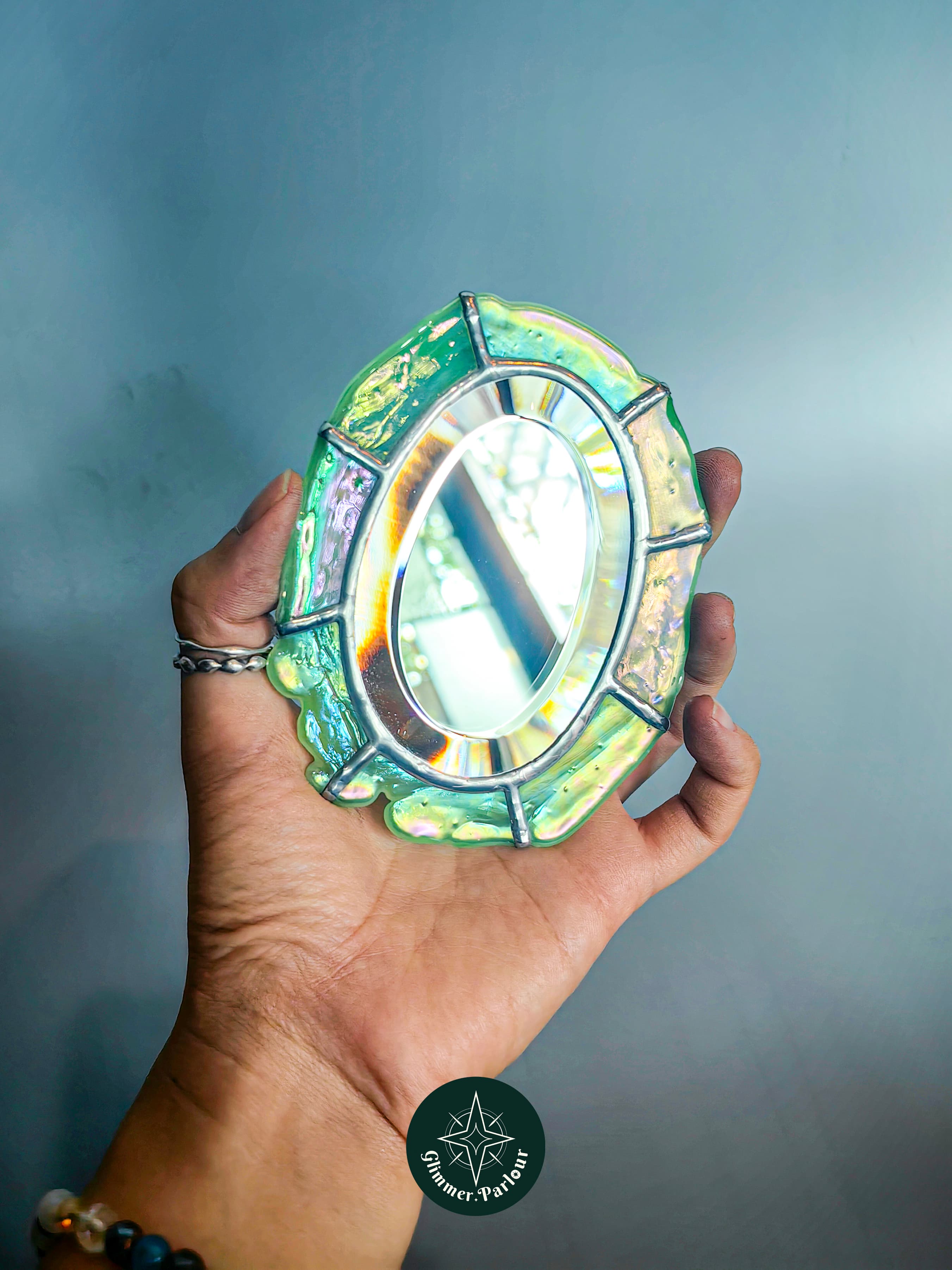 Hand holding a small, oval mirror framed with iridescent, light green stained glass.
