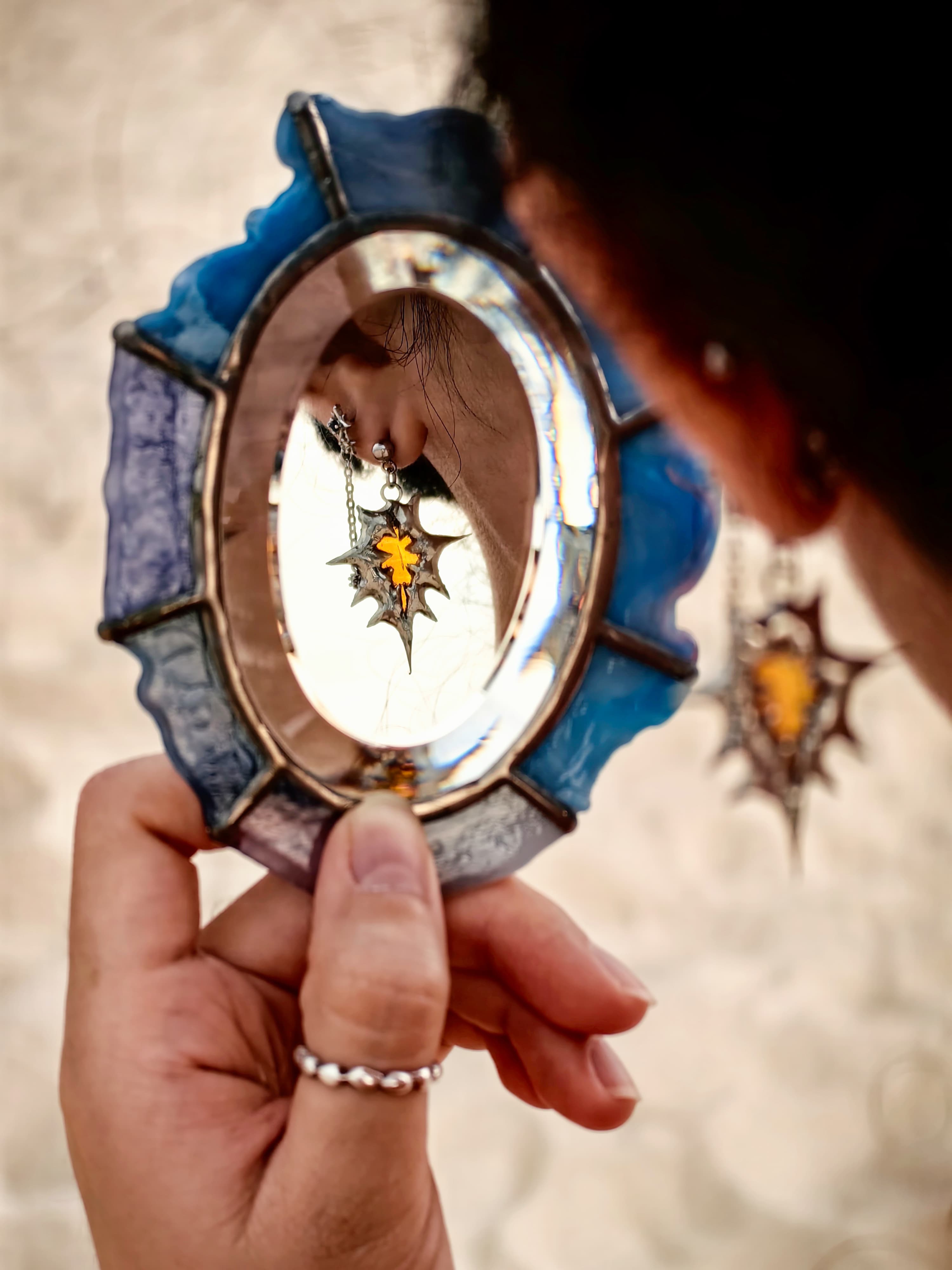 Hand holding a blue mirror reflecting a large star-shaped earring with an amber center.