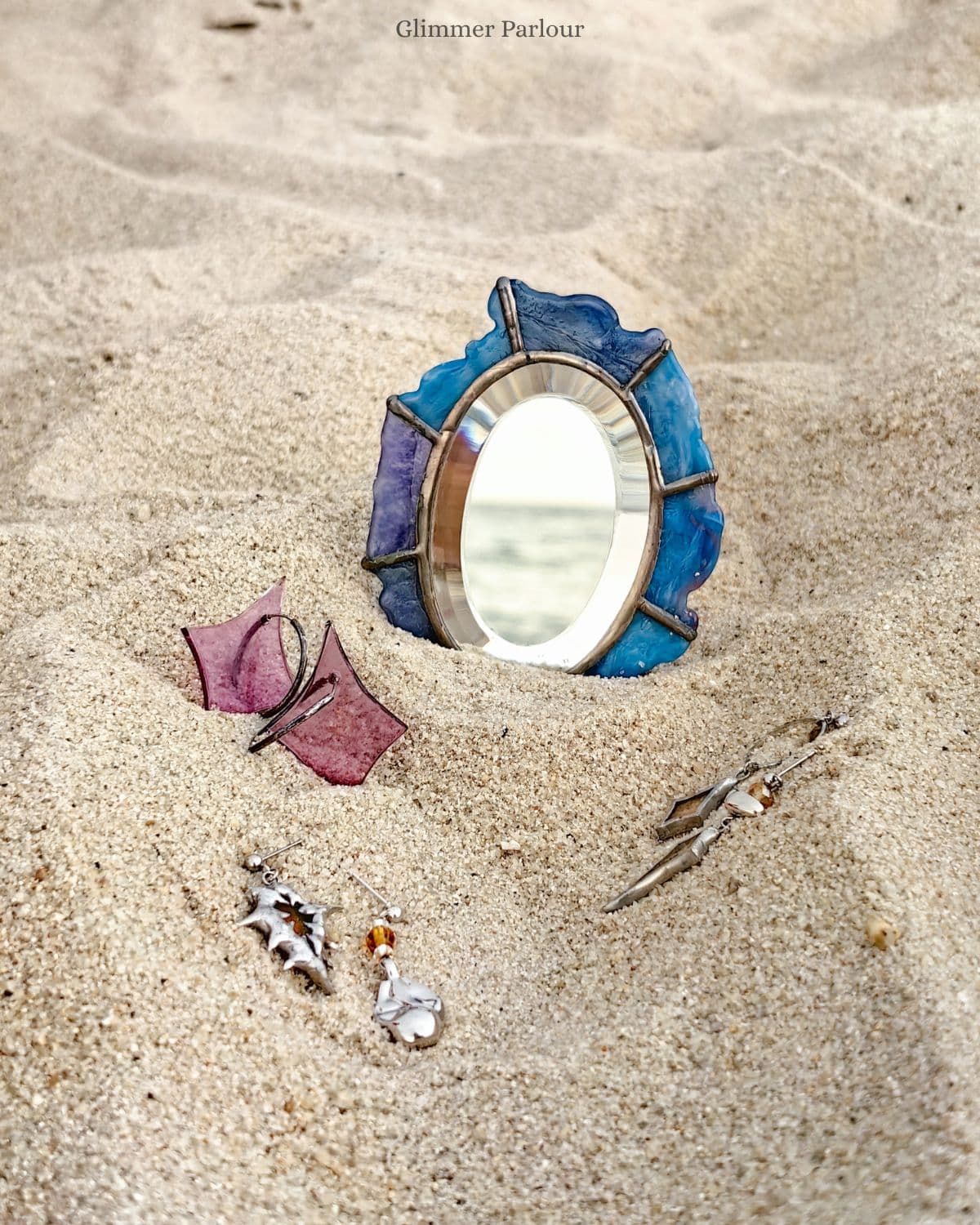 Blue stained glass mirror reflecting the sea, with jewelry scattered on a sandy beach.