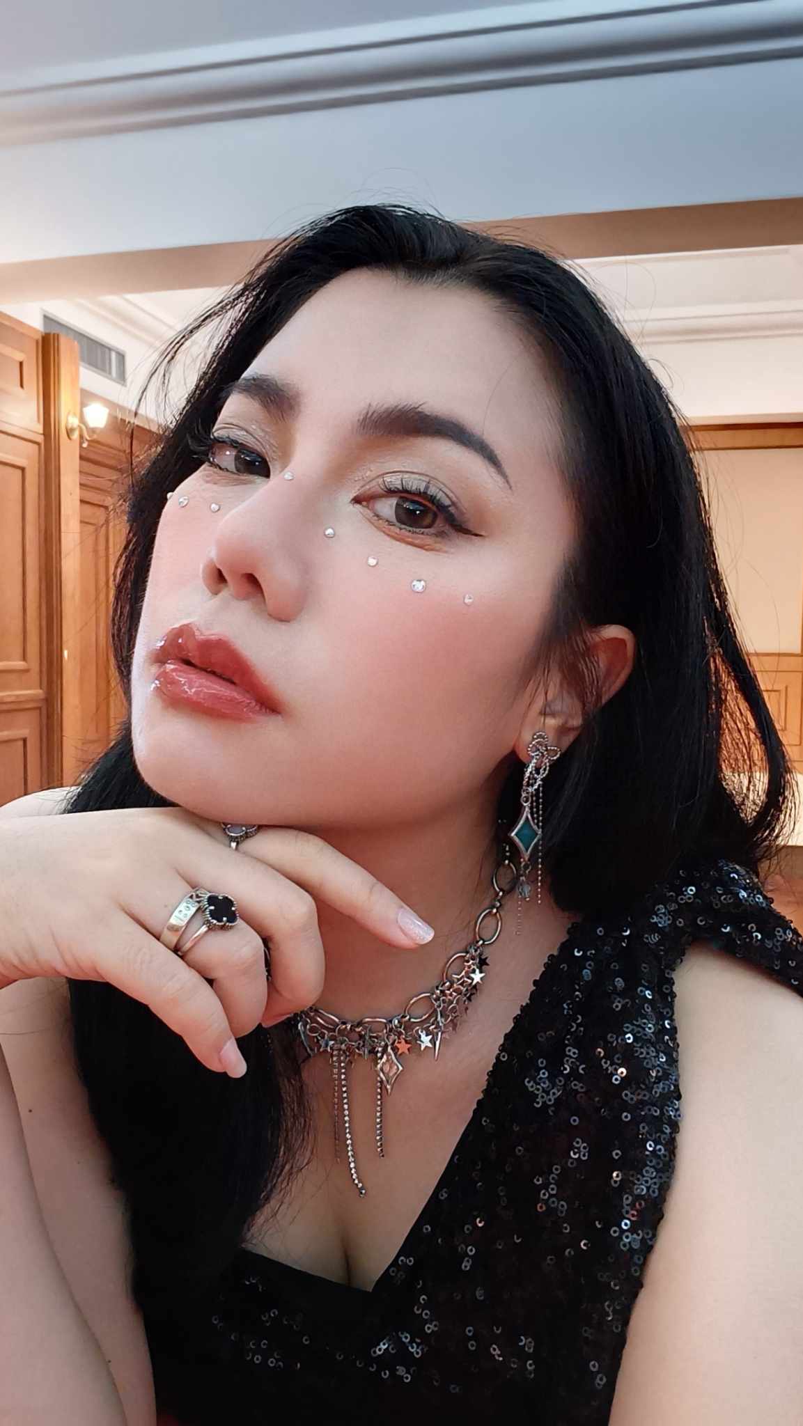 Woman with face gems and silver jewelry posing in a black sequined top, hand under chin.