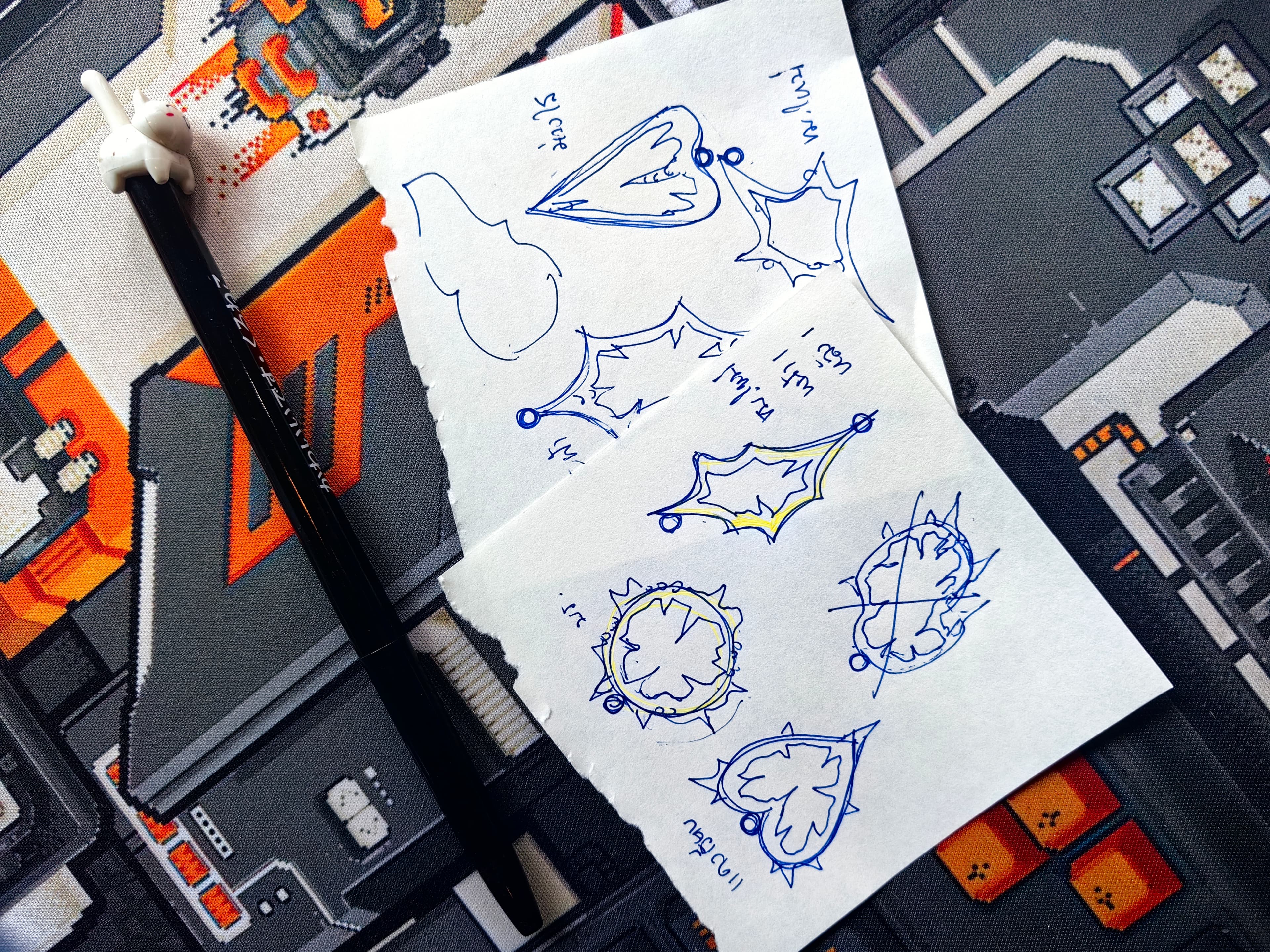 Jewelry design sketches on paper scraps beside a cat pen on a pixel art mat.